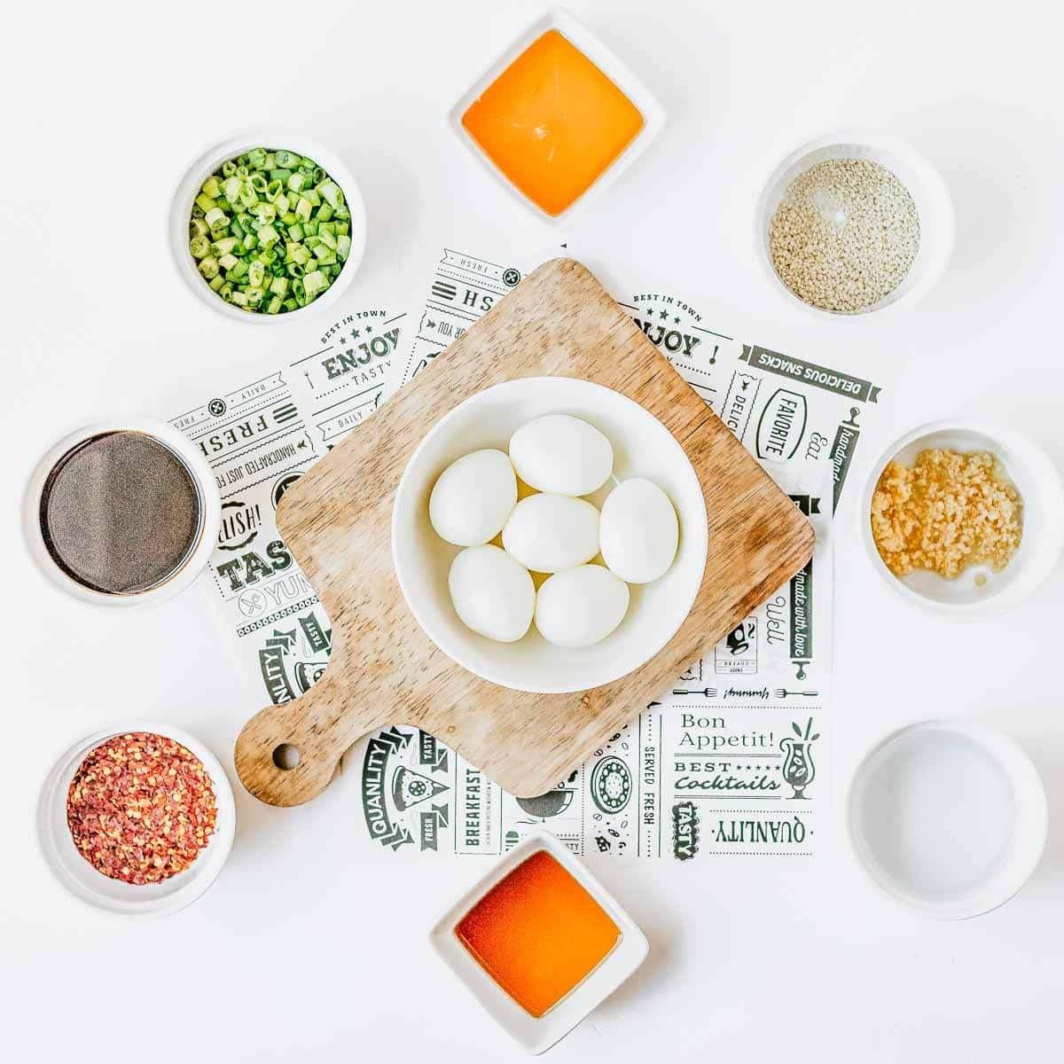 Overhead view of a cutting board with six boiled eggs in a bowl, surrounded by small bowls of chopped green onions, sesame seeds, minced garlic, black pepper, red pepper flakes, and two orange sauces on a white background.