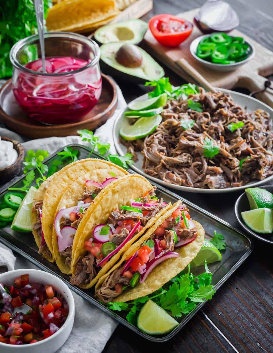 Lamb taco spread on a table with lime wedges, braised lamb meat, pico de gallo, crema, jalapenos, avocado and pickled radishes and red onion.