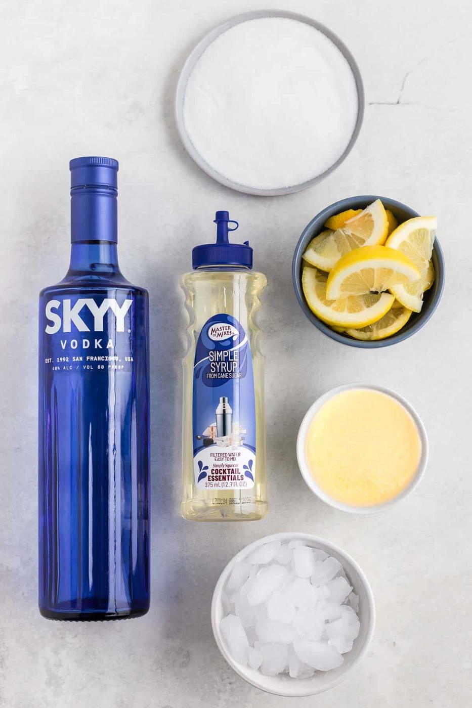 Overhead view of cocktail ingredients: a blue bottle of Skyy vodka, a bottle of simple syrup, a bowl of sugar, a bowl of lemon slices, a bowl of lemon juice, and a bowl of ice on a light surface.