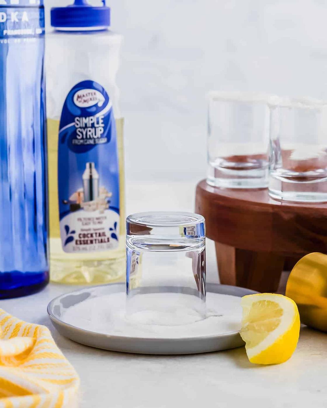 A glass rim being dipped in sugar on a plate. Nearby are simple syrup, a bottle, shot glasses on a wooden stand, a lemon wedge, and a yellow-striped cloth. The setup suggests preparation for making cocktails.