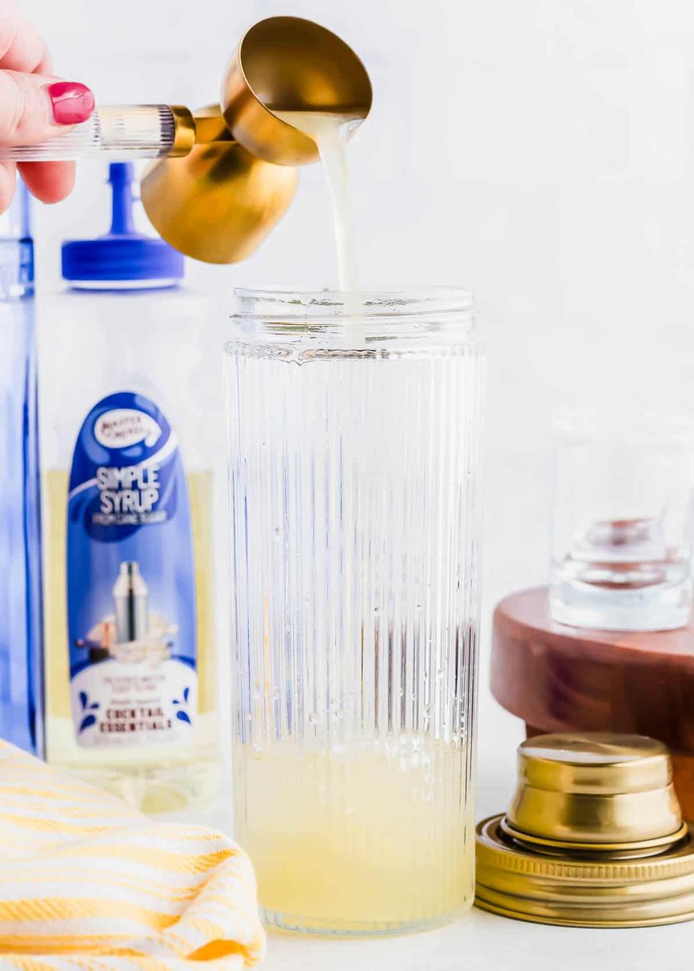 A hand pours liquid from a brass measuring cup into a tall, ridged glass. Behind the glass, there's a bottle labeled "Simple Syrup" and a wooden coaster. A yellow-striped cloth and a gold lid are nearby.