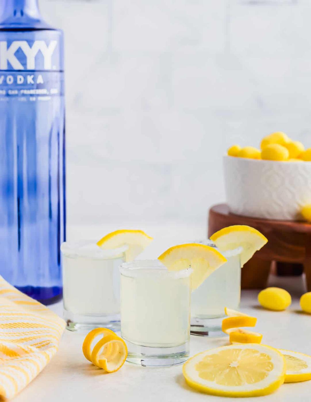 Three lemon drop cocktails in shot glasses, garnished with lemon slices, sit on a white surface. A blue vodka bottle and a bowl of yellow candies are in the background. Lemon wedges are scattered around, along with a yellow striped towel.
