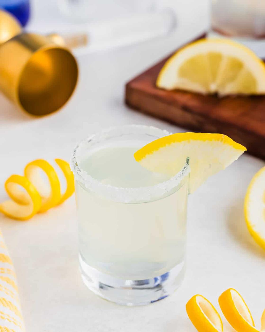 A salt-rimmed glass with a lemon wedge garnish contains a lemon drop shot. Curled lemon peels and sliced lemons are scattered around on the white surface. A wooden board, golden jigger, and cloth are visible in the background.