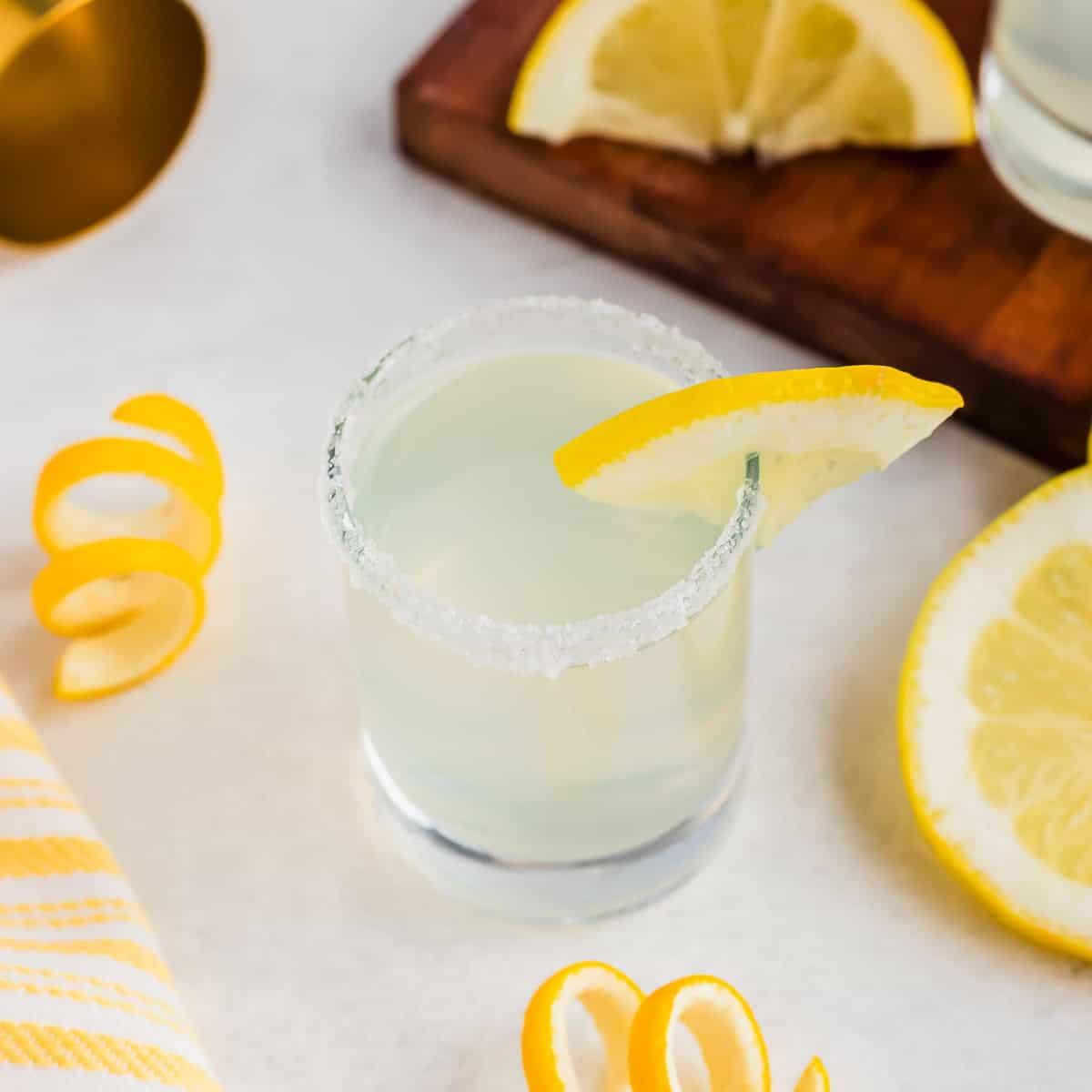 A glass of lemon drop shot cocktail with a sugared rim and lemon wedge garnish sits on a white surface. Nearby are lemon slices and peels, a yellow-striped cloth, and a wooden board with more lemon slices.