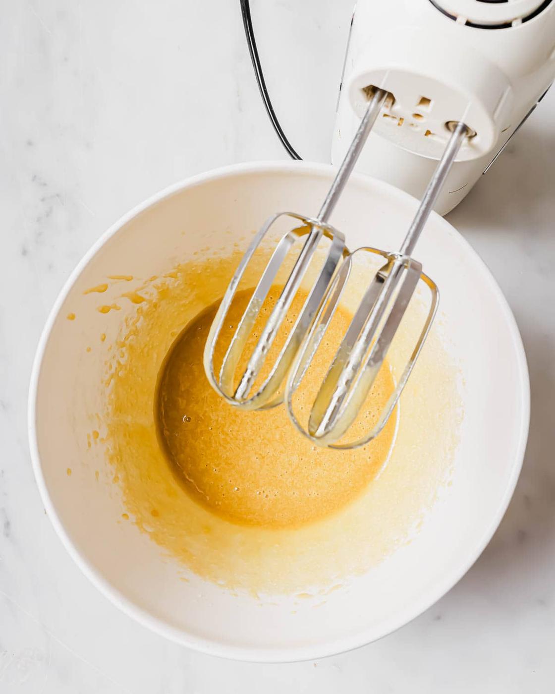 A white mixing bowl containing a yellow batter, positioned on a marble countertop. An electric hand mixer with two metal beaters rests over the bowl.