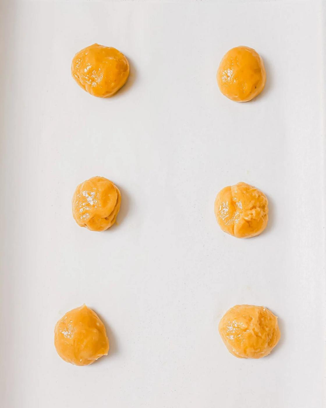 Six small, round, cookie dough balls are evenly spaced on parchment paper, ready for baking.