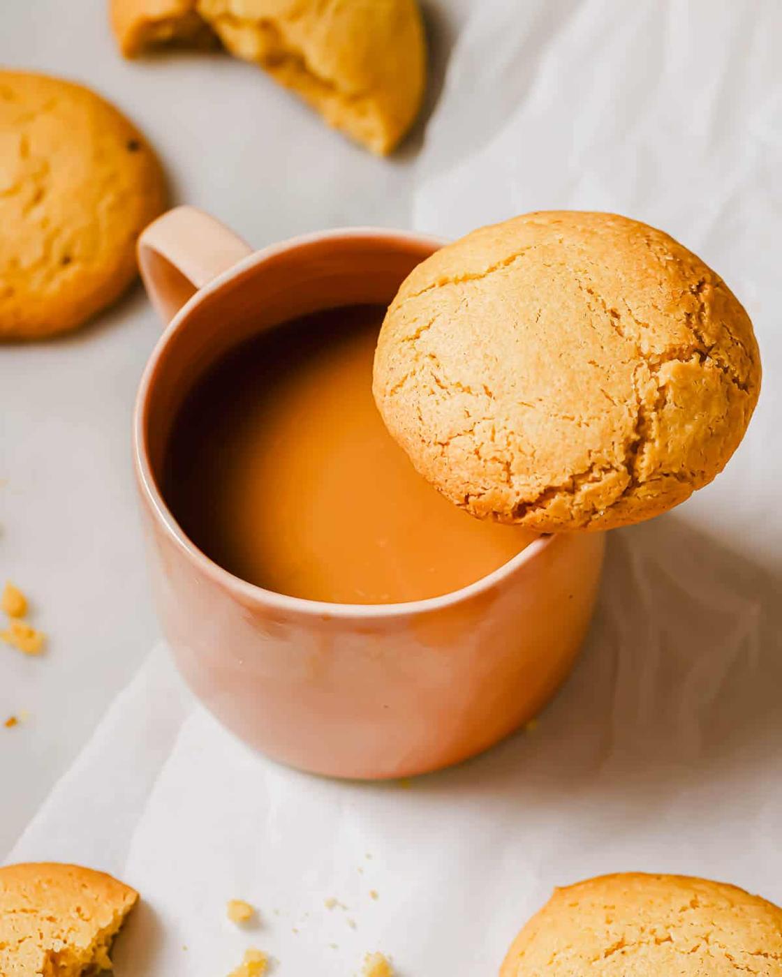 A pink mug filled with coffee is on a white surface. A crinkle olive oil cookie rests on the rim, and several other cookies are nearby, with some crumbs scattered around.
