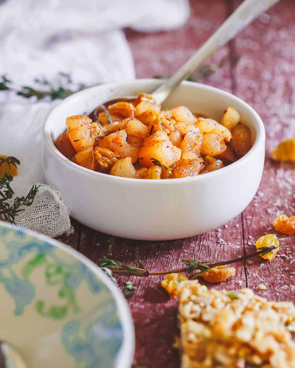 A bowl of cooked apple compote seasoned with herbs and spices, with a spoon in the bowl. Pieces of herbs and crumbs are scattered on a wooden surface nearby.