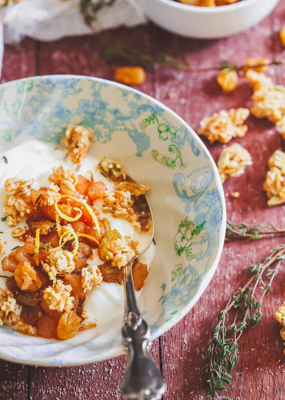 A bowl of yogurt topped with apple compote, granola and citrus zest, placed on a wooden table next to herbs and scattered granola pieces. A spoon rests inside the bowl.