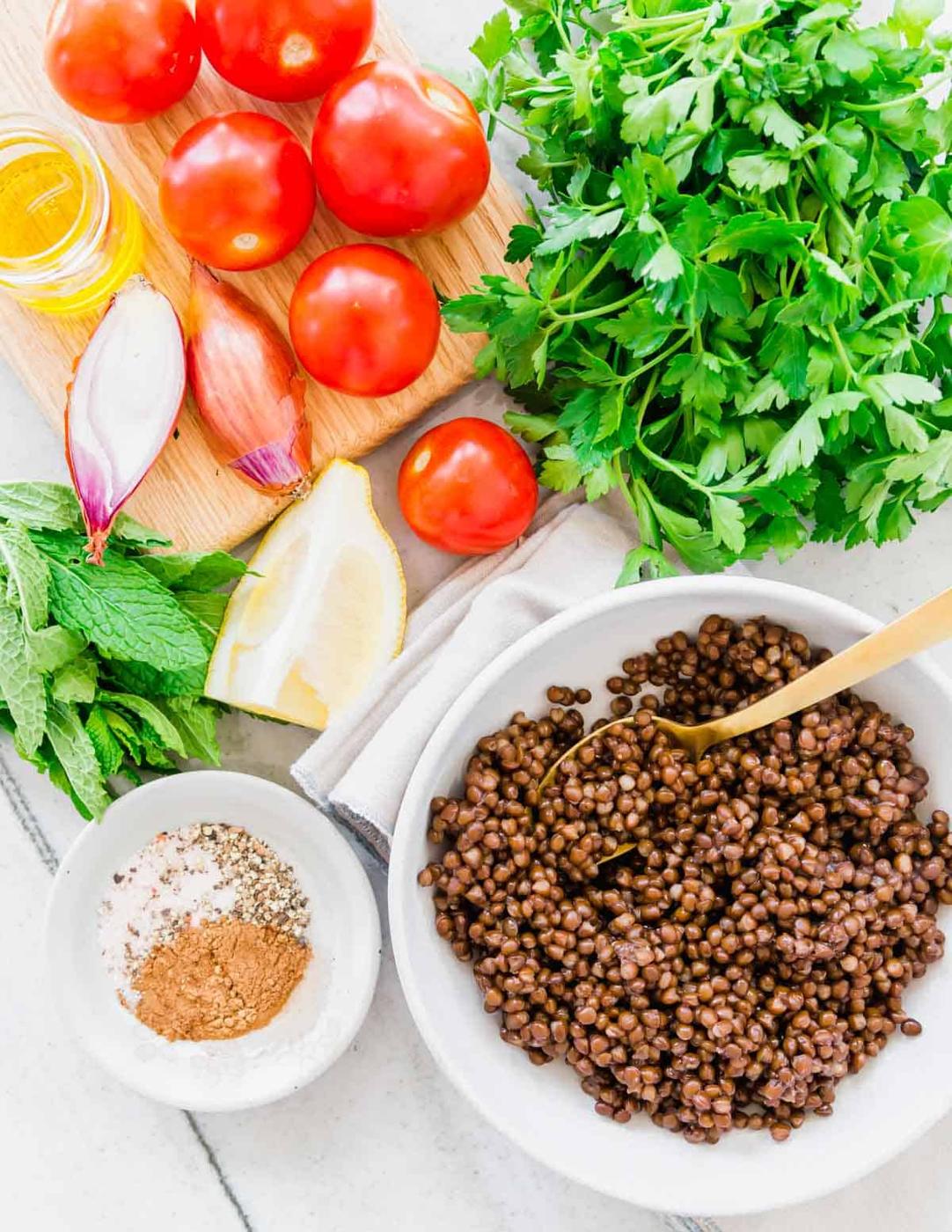 Cooked lentils in a bowl with gold spoon with ingredients to make tabbouleh around it.