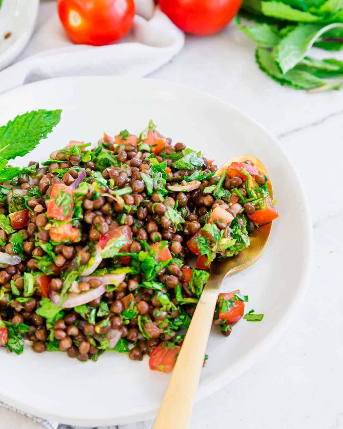 Gluten-free lentil tabouli recipe with herbs, shallots, tomatoes and a lemon spice dressing on a white plate with gold spoon.