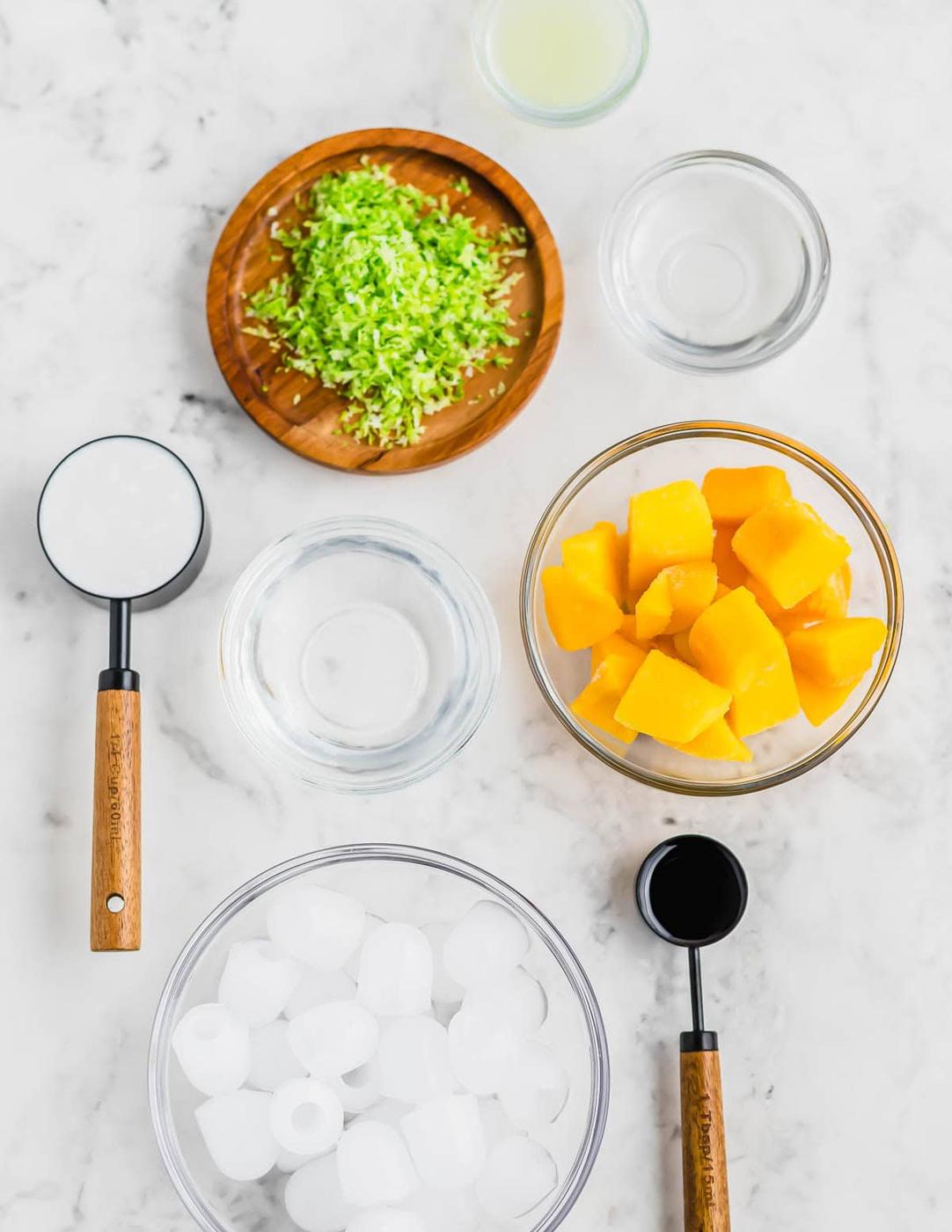 Top view of a marble surface with ingredients for a margarita recipe including diced mango in a glass bowl, chopped green herbs on a wooden plate, a bowl of ice cubes, and measuring cups with liquid. Some empty bowls are also present.