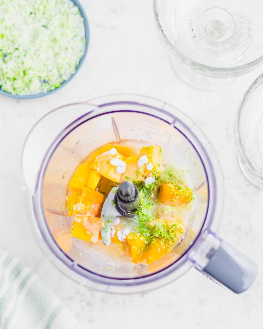 Top view of a blender containing chunks of mango, lime zest, and coconut milk. A small bowl of lime zest and sugar and two empty glasses are nearby on a white countertop. A striped cloth is partially visible in the corner.