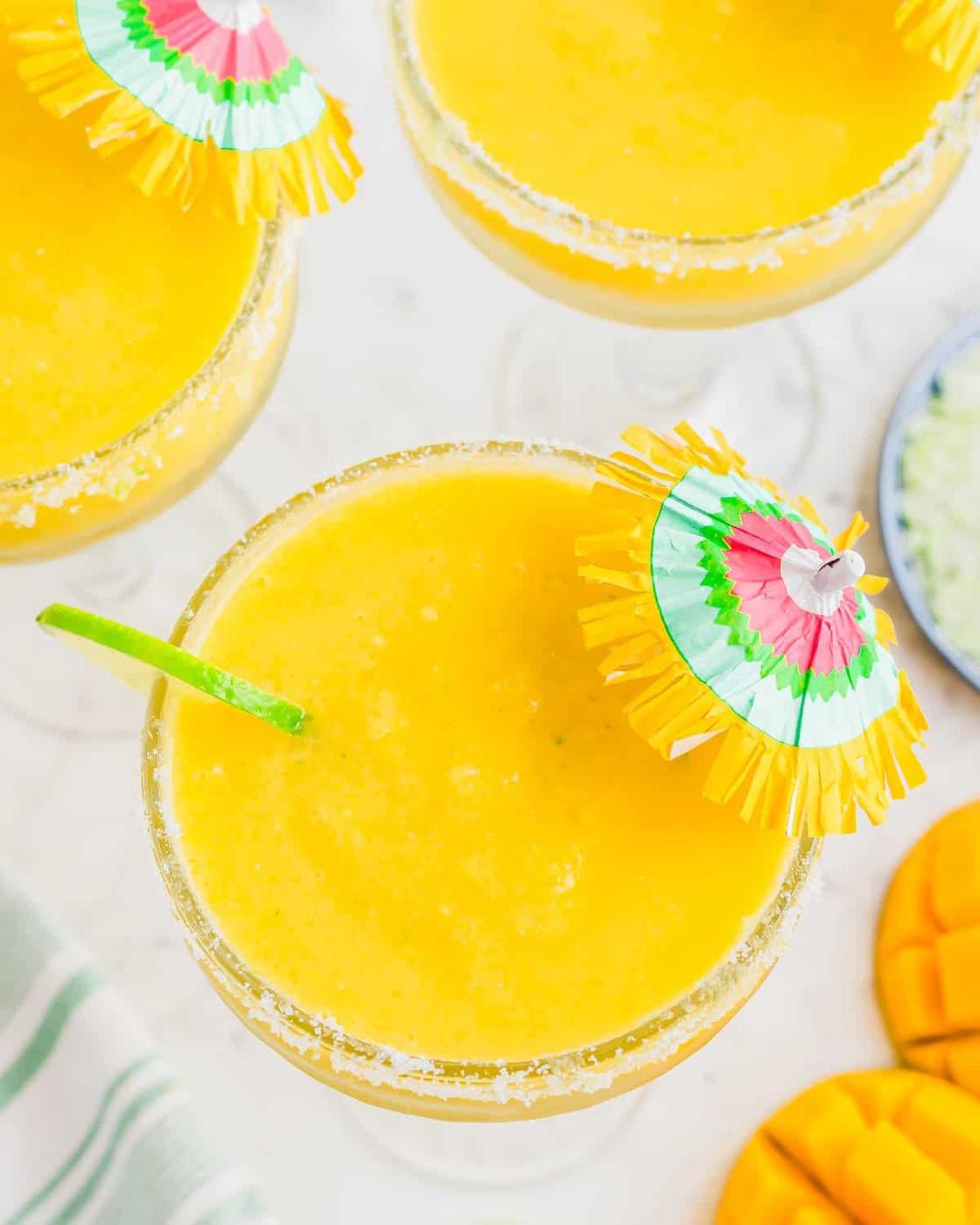 Top view of vibrant yellow mango margaritas in glasses with lime and sugar rims. Each drink is garnished with a colorful paper umbrella and a lime wedge. Slices of fresh mango are nearby, and a striped cloth is partially visible.