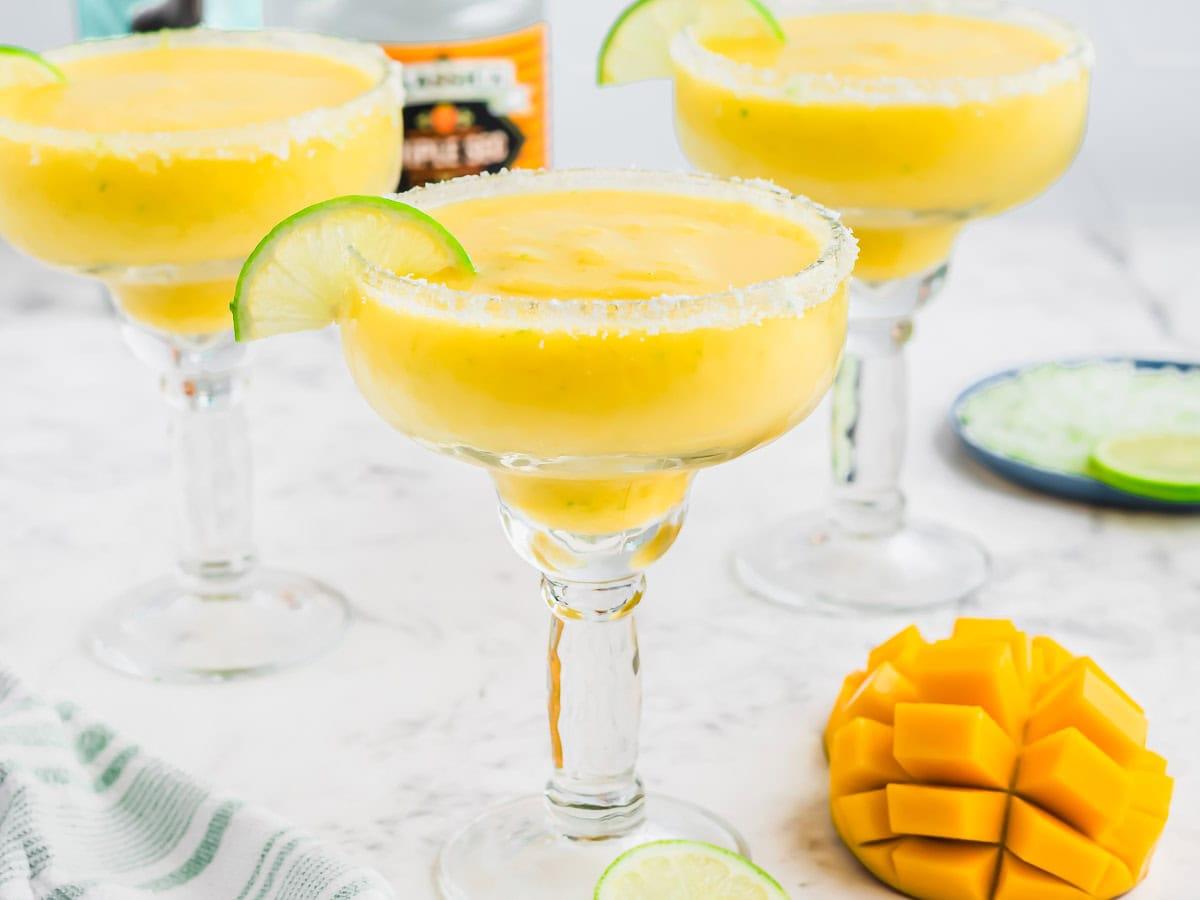 Three margarita glasses filled with frozen mango margaritas, garnished with lime slices and salt on the rims, are placed on a marble surface. A diced mango and more lime slices are nearby.
