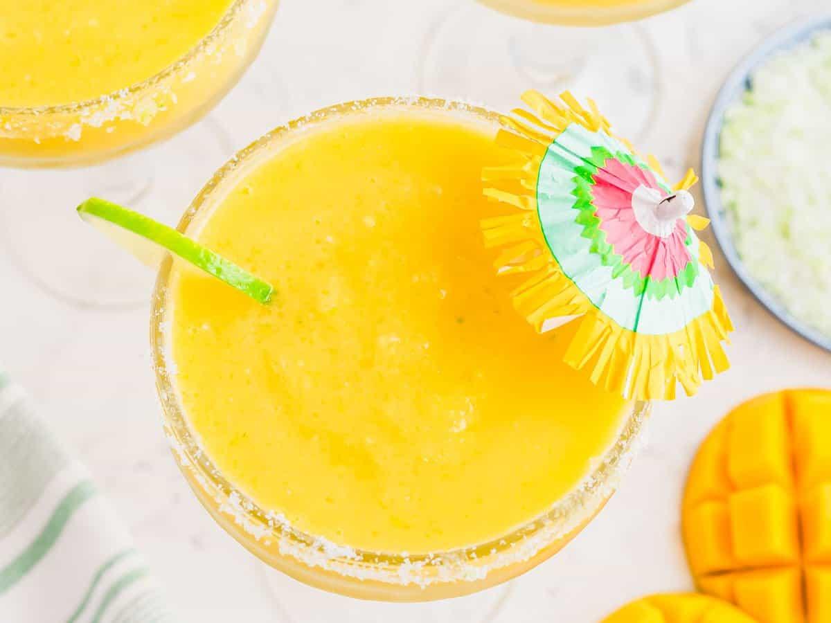 Top view of a vibrant yellow mango margarita in a glass with a sugared rim, garnished with a lime slice and a colorful cocktail umbrella. A sliced mango and a plate with salt can be seen in the background.