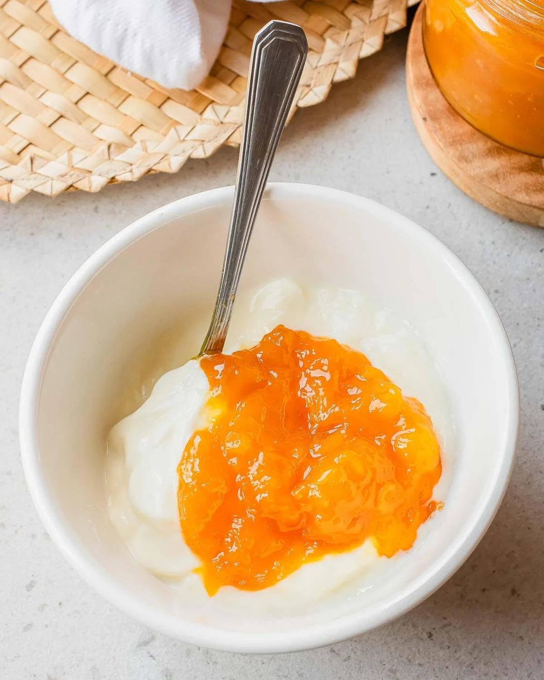 A white bowl filled with plain yogurt topped with bright orange mango jam, with a spoon inside. The bowl sits on a light countertop near a woven mat and a jar of orange jam.