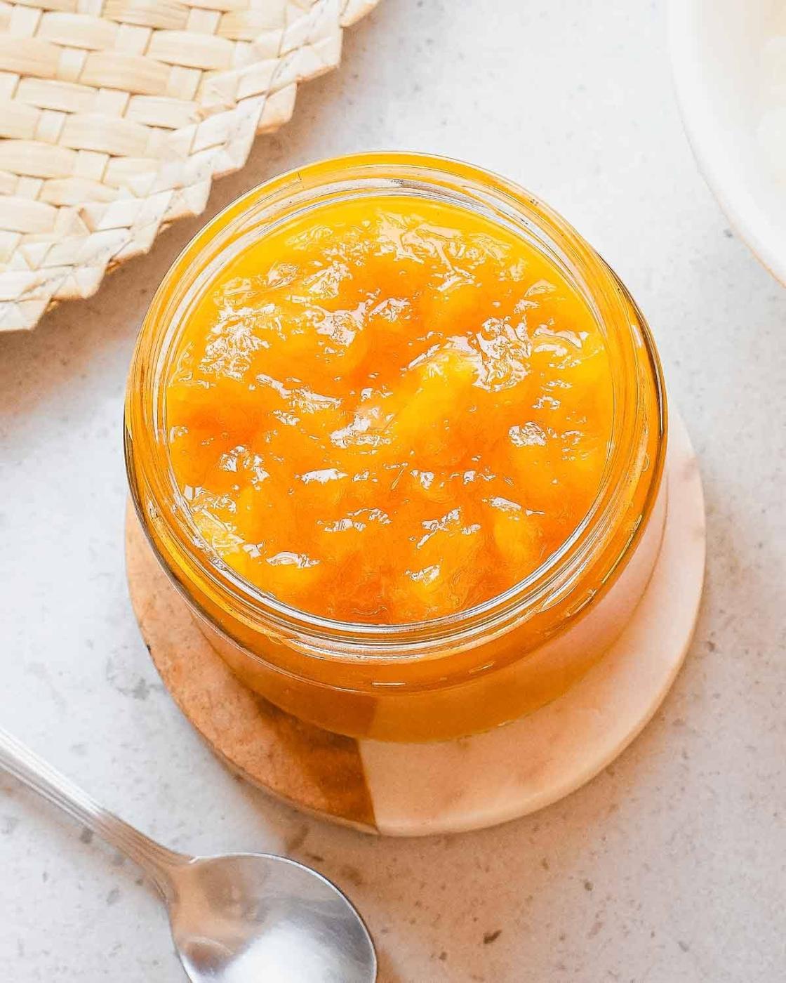 A glass jar filled with bright orange homemade mango jam sits on a round coaster, next to a spoon and a woven placemat on a light-colored surface.
