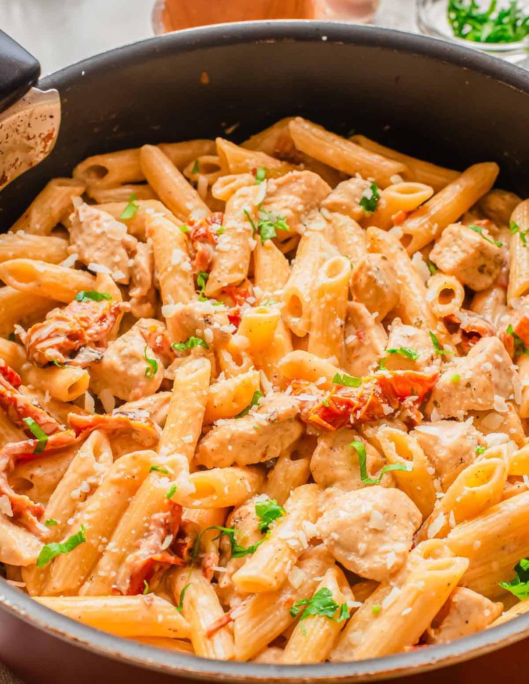 A skillet filled with creamy penne pasta, chunks of chicken, sun-dried tomatoes, and garnished with chopped parsley and grated cheese.