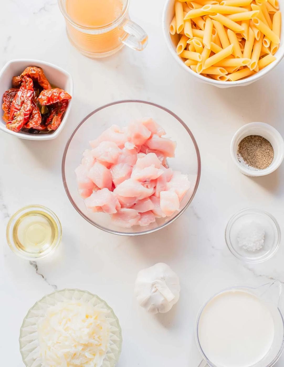 Overhead view of neatly arranged ingredients: cubed raw chicken, penne pasta, grated cheese, sun-dried tomatoes, garlic, cream, broth, oil, salt, and pepper on a white marble surface.