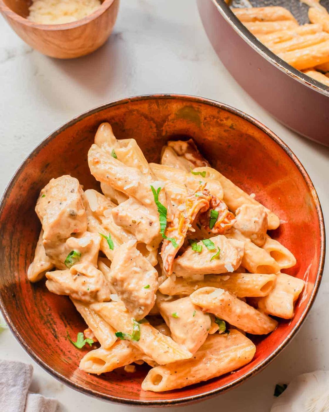 A bowl of creamy penne pasta with pieces of chicken, garnished with chopped herbs. A skillet and a small bowl of grated cheese are visible in the background.