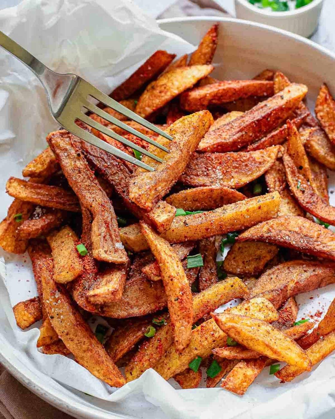 A plate of crispy, seasoned homemade masala fries garnished with chopped herbs, with a fork picking up a few pieces. The fries rest on white parchment paper inside a round dish.