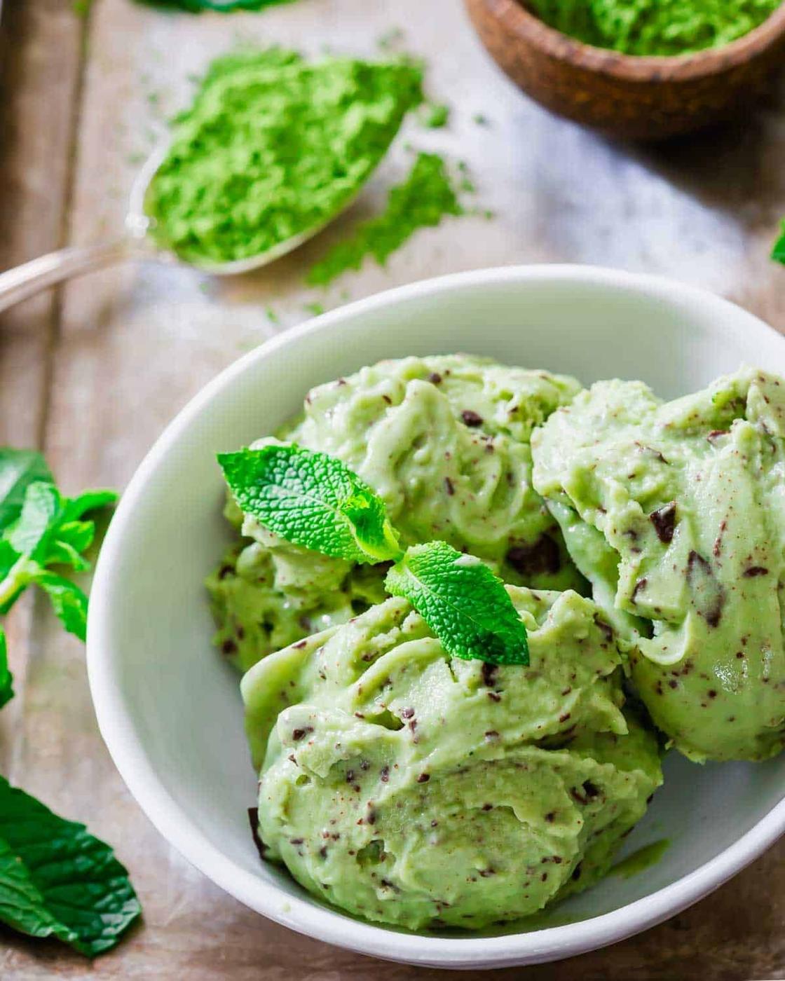 A bowl of matcha ice cream garnished with fresh mint leaves. Scoops are creamy with visible chocolate flakes. A spoonful of powdered matcha is in the background, suggesting a flavor combination with the ice cream.