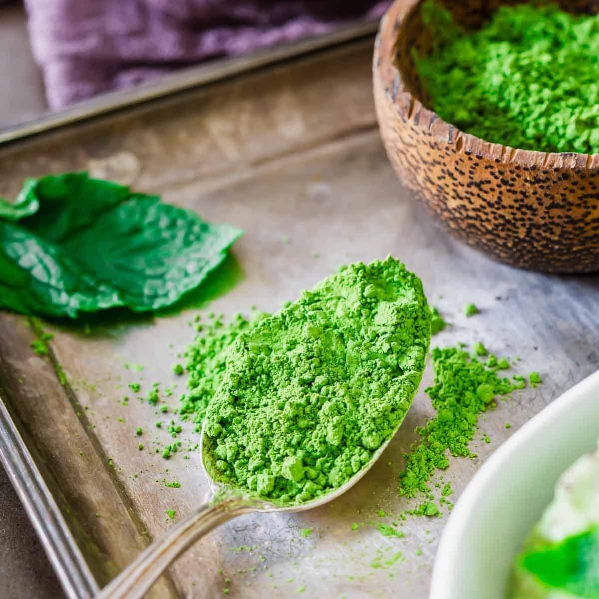 A spoon filled with vibrant green matcha powder rests on a metal tray. A wooden bowl with more matcha is in the background, and a green leaf lies beside the spoon. The scene conveys a natural, organic feel.