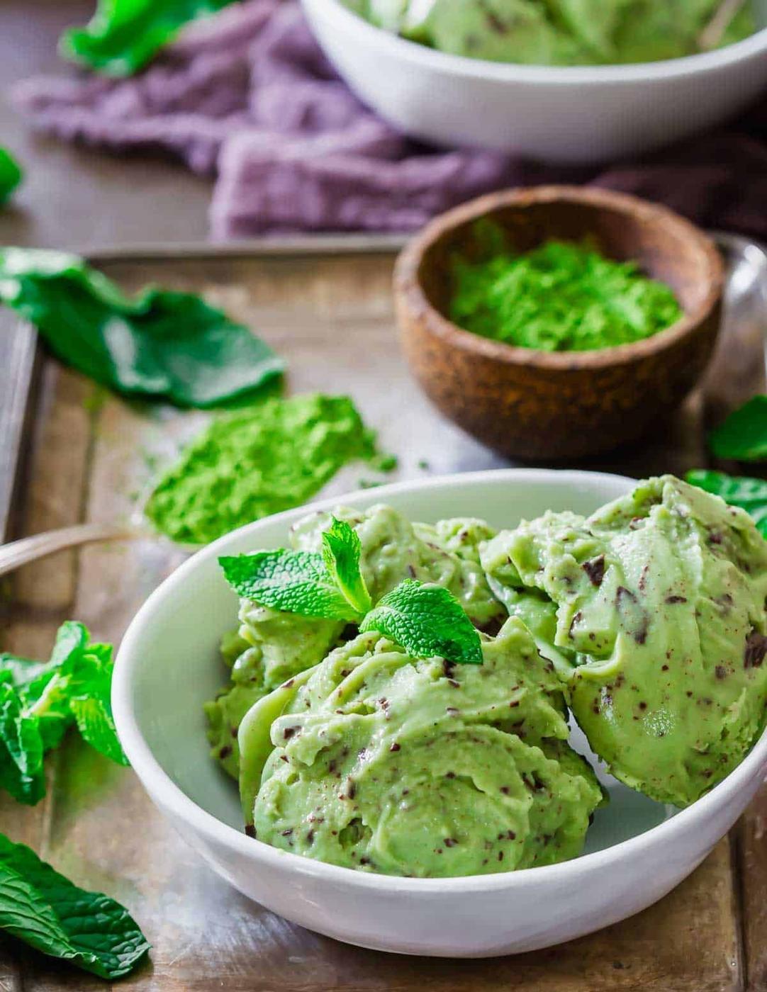 A white bowl filled with matcha mint chocolate chip ice cream is garnished with fresh mint leaves. A wooden bowl with green matcha powder and more mint leaves are in the background on a wooden tray.