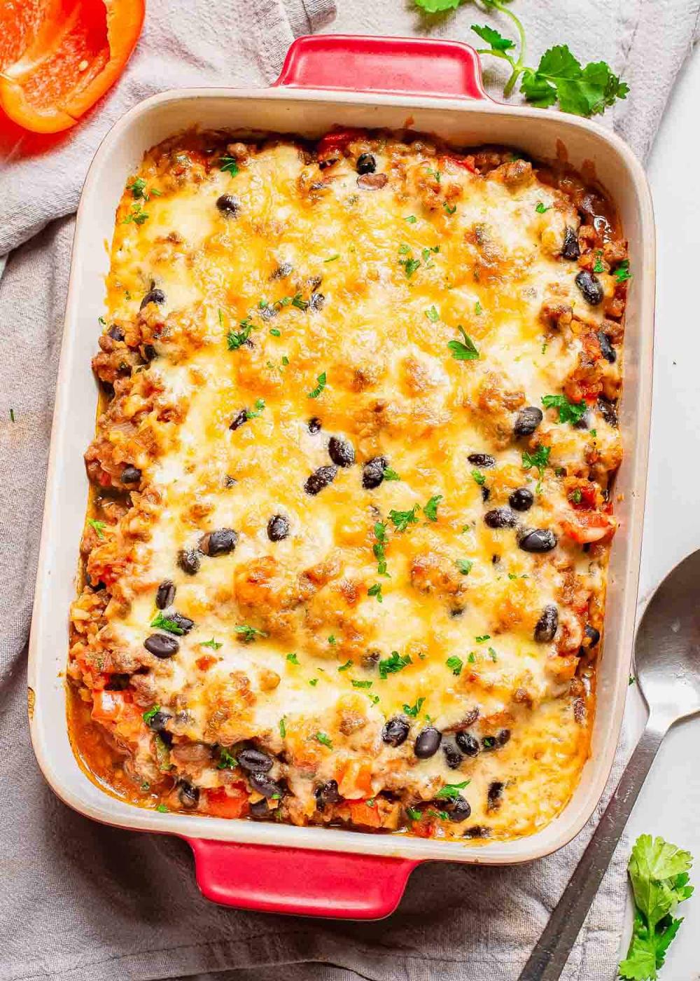 A baked casserole dish filled with melted cheese, ground meat, black beans, and chopped vegetables, garnished with herbs, is placed on a cloth with a spoon beside it.