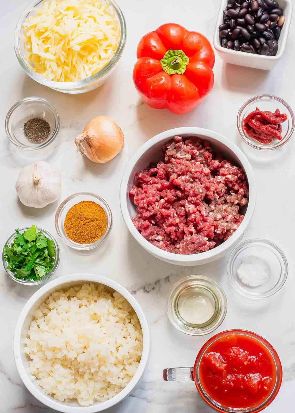 Ingredients for a casserole: ground beef, red bell pepper, black beans, grated cheese, cauliflower rice, garlic, onion, parsley, spices, tomato sauce, and small bowls of seasonings on a marble surface.