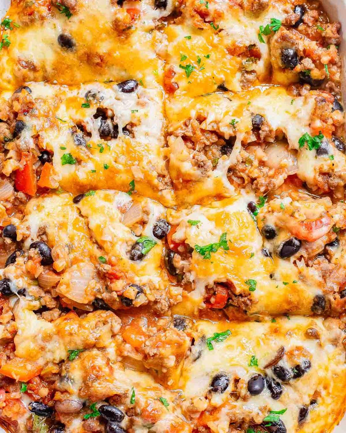 A close-up of a casserole featuring melted cheese, ground meat, black beans, and diced tomatoes, garnished with chopped parsley.