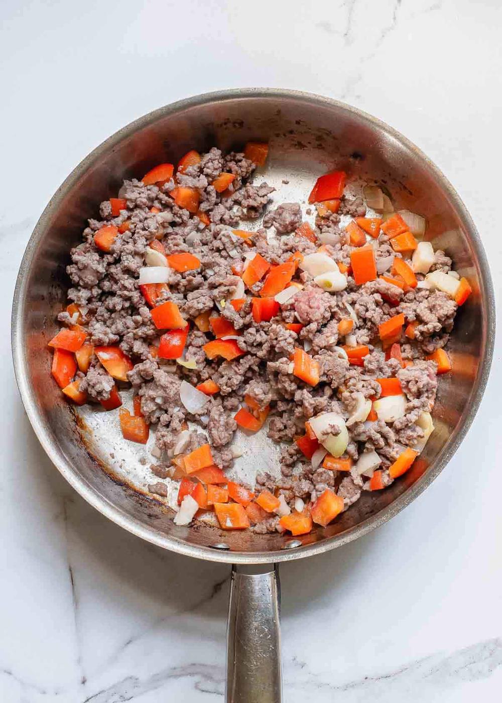 Ground beef cooked with chopped red bell peppers and onions in a frying pan on a marble surface.