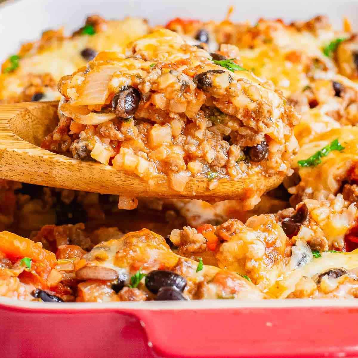 A wooden spoon scoops a cheesy, baked beef and rice dish with black beans and vegetables from a red casserole dish.
