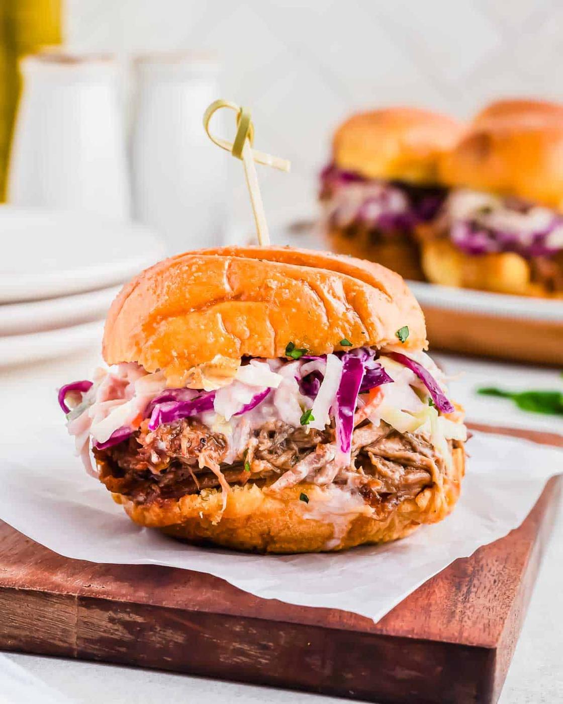 A pulled pork slider sandwich topped with coleslaw is placed on a wooden serving board with a skewer through the top; more sandwiches are visible in the background.