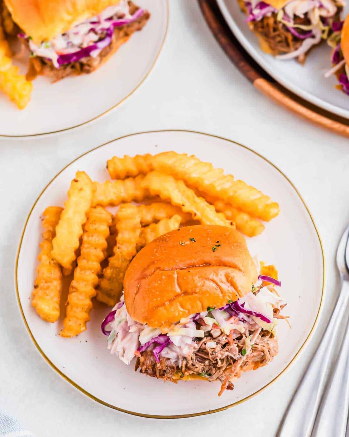 A plate with a pulled pork sandwich topped with coleslaw, accompanied by crinkle-cut fries. Additional sandwiches are visible in the background.