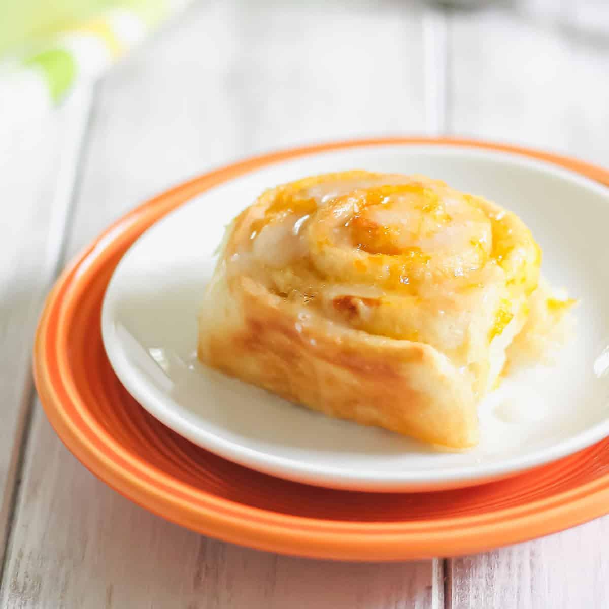 An orange glazed cinnamon roll is placed on a white plate with an orange rim, set on a light wooden surface.