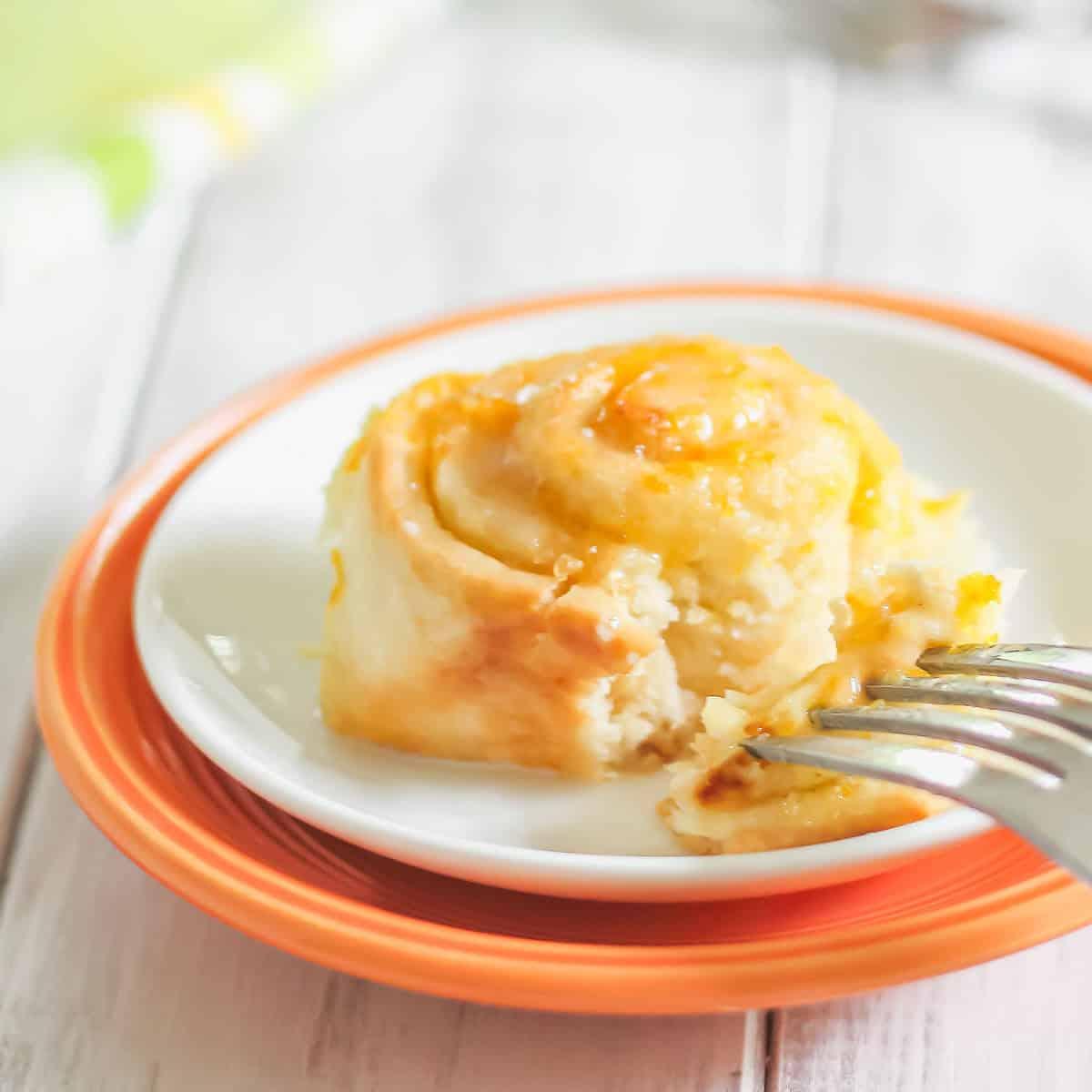 A close-up of a cinnamon roll with orange jam icing on a white plate with an orange rim. A fork is cutting into the cinnamon roll.