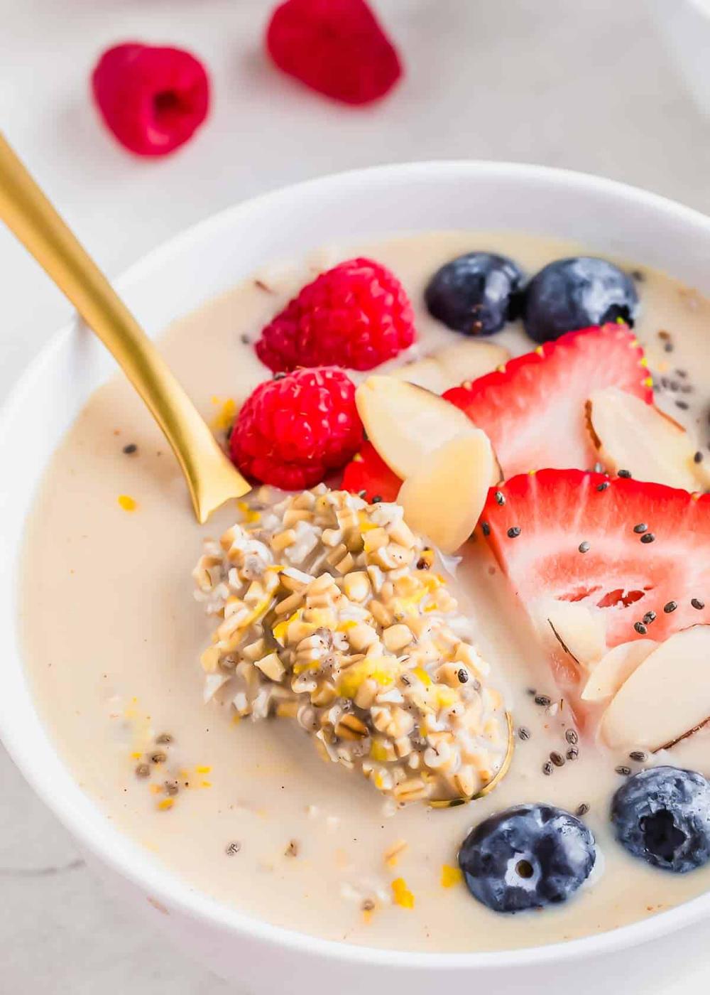 A gold spoon lifting overnight steel cut oats out of a serving bowl. The bowl also contains fresh berries and almond slices.