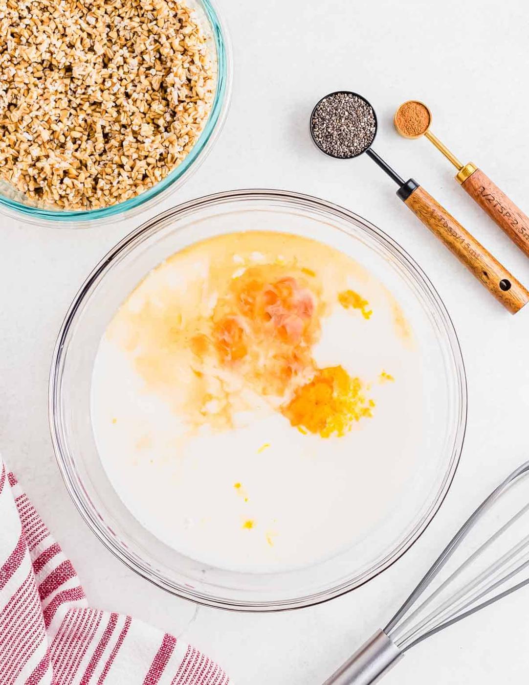 A bowl with milk, orange juice, vanilla and orange zest. Soaked steel cut oats sit in another bowl to the side with two small measuring spoons containing chia seeds and cinnamon. There's a whisk and kitchen towel slightly out of frame.