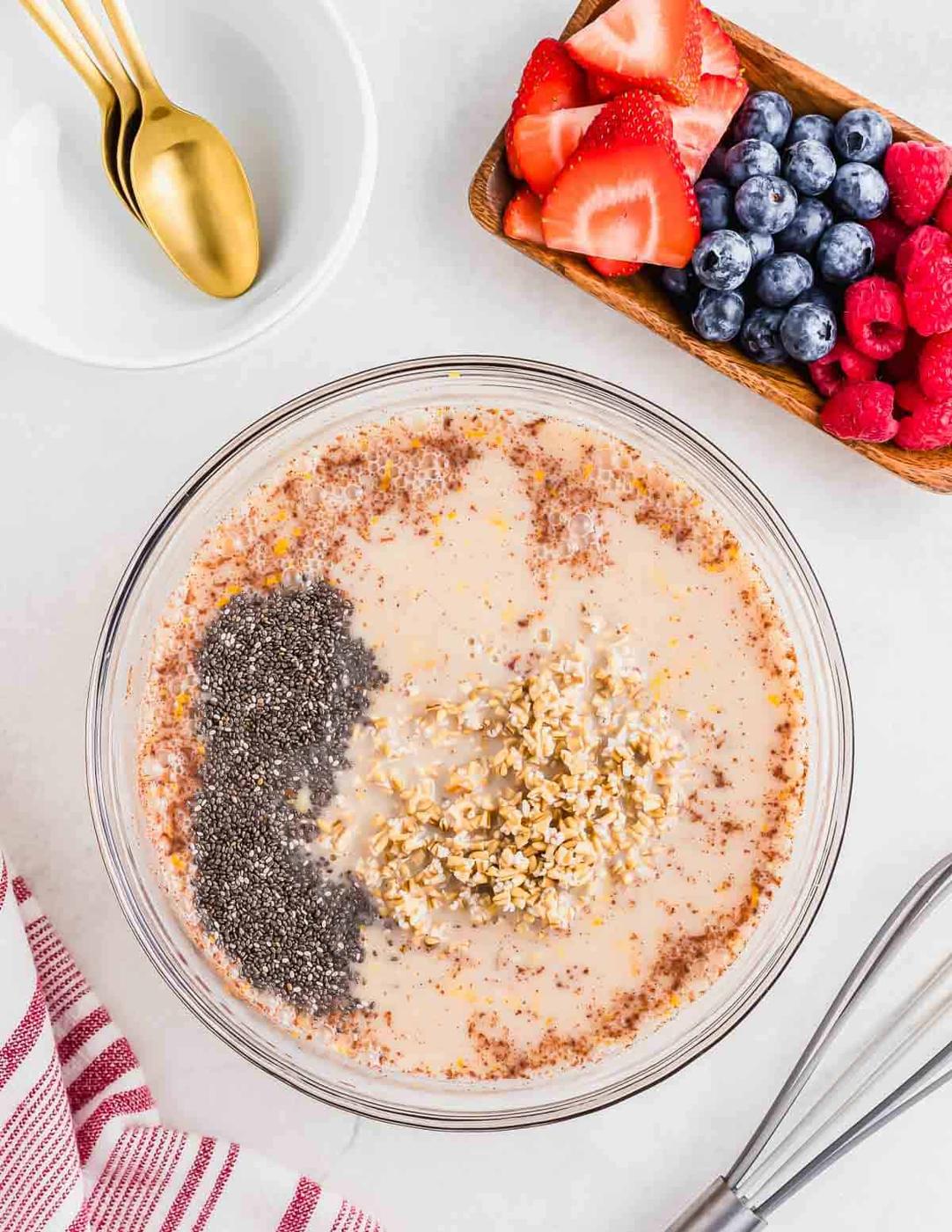 All the ingredients to make overnight steel cut oats in a large glass bowl. There are berries on the side with a whisk and a few white bowls with gold spoons.