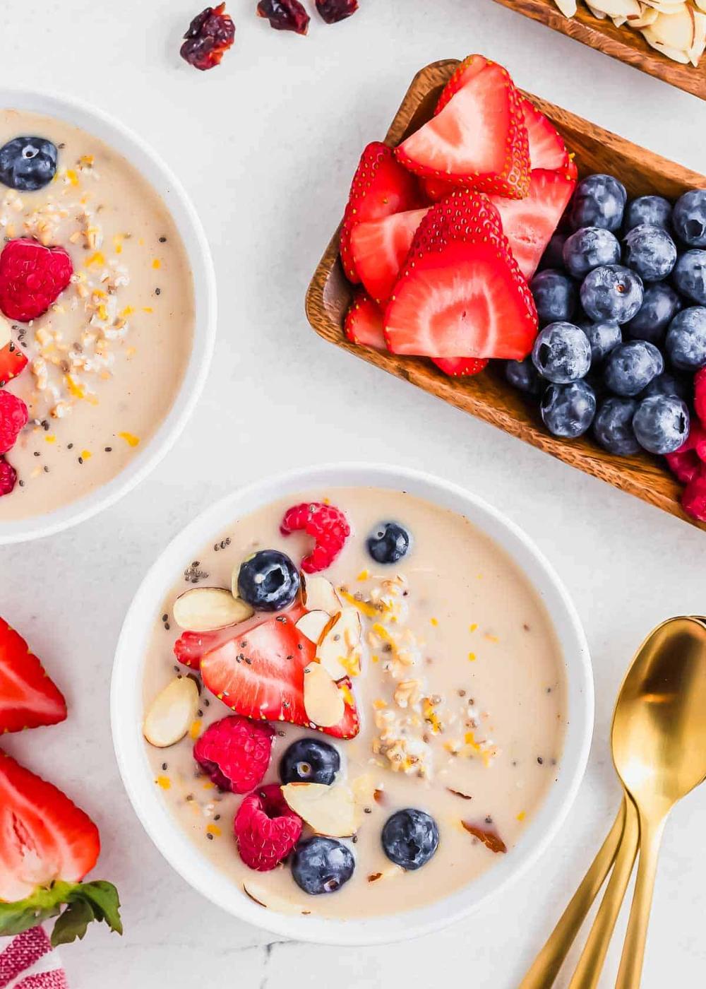 Overnight steel cut oats in a white bowl topped with fresh berries and sliced almonds. Gold spoons are on the side and more berries on a tray.