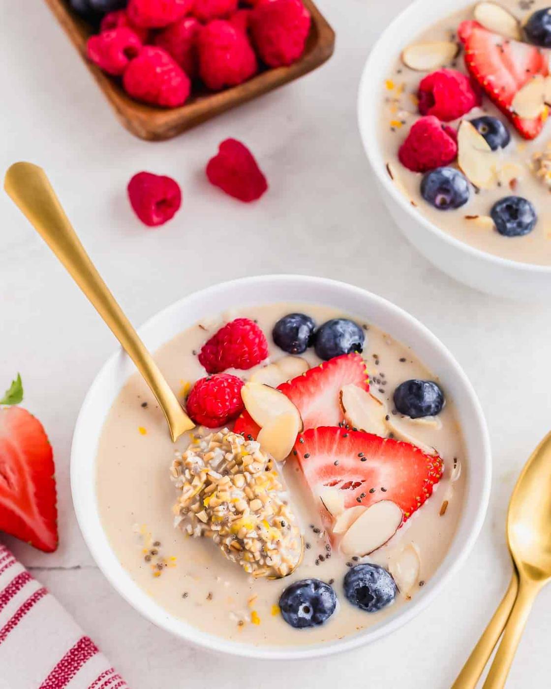 Overnight oats made with steel cut oats in a white serving bowl with a gold spoon lifting out a spoonful from the bowl. Berries are scattered about and a few more spoons sit to the side.