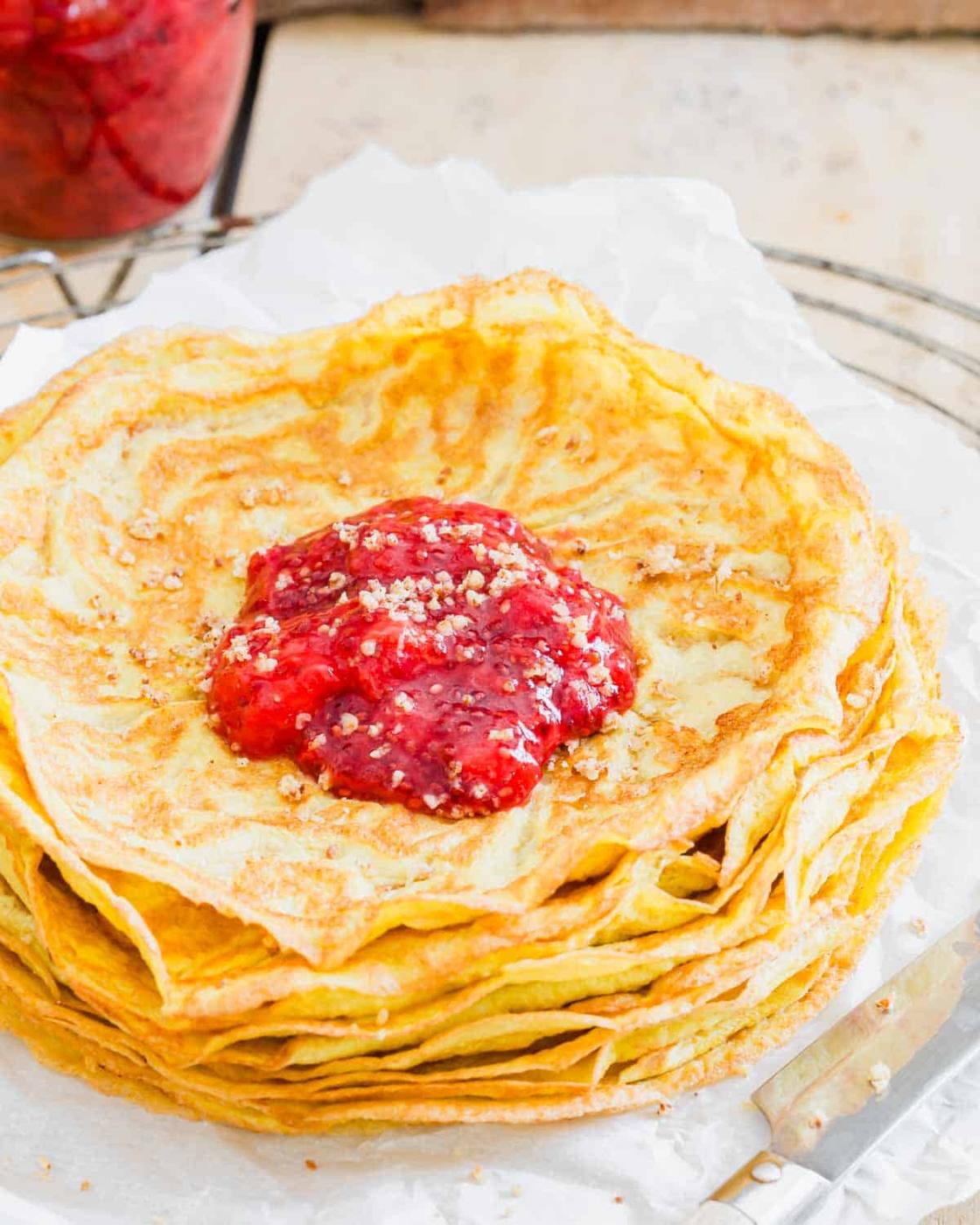 A stack of thin, golden paleo crepes topped with a dollop of red berry compote and sprinkled with powdered sugar, served on a white paper sheet.