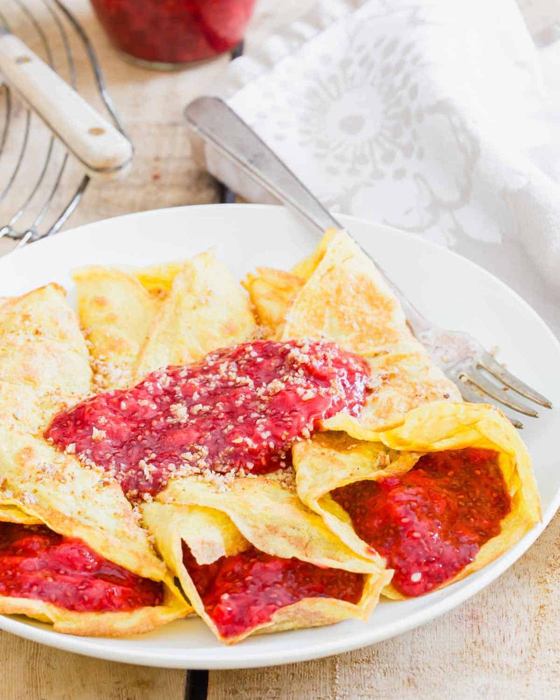 A plate of folded grain-free crepes topped with red strawberry sauce and powdered sugar, served on a rustic wooden table.