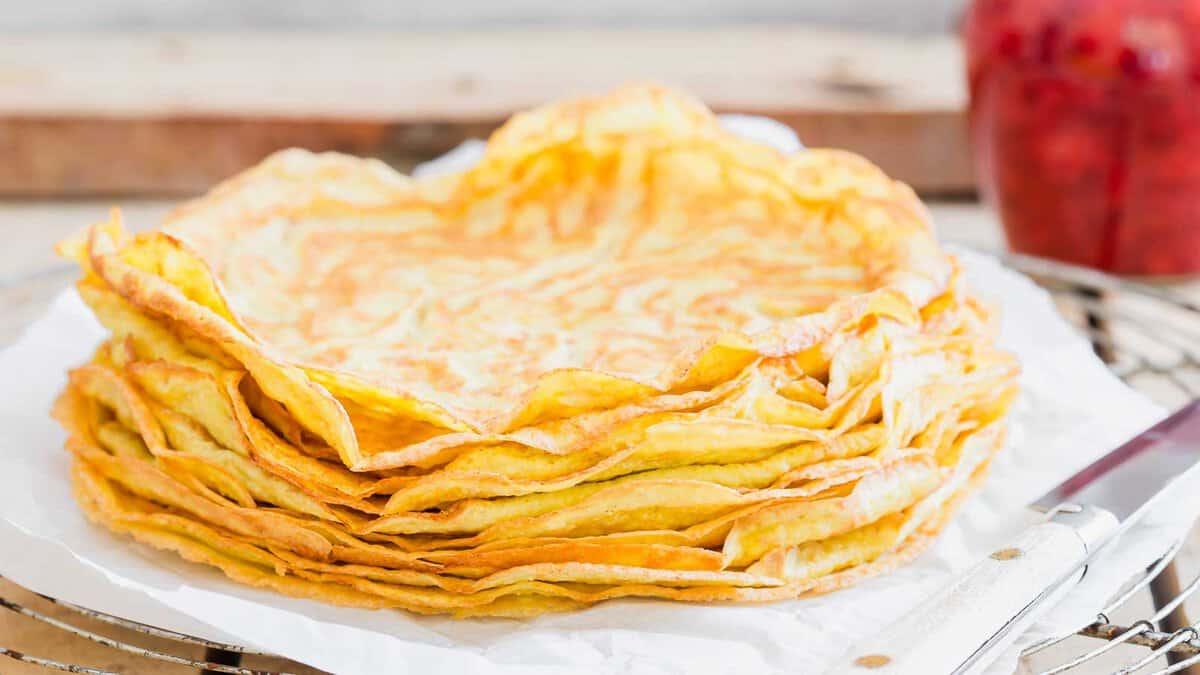 A stack of golden, crispy crepes on a white paper, with a jar of jam and a knife in the background on a wooden table.