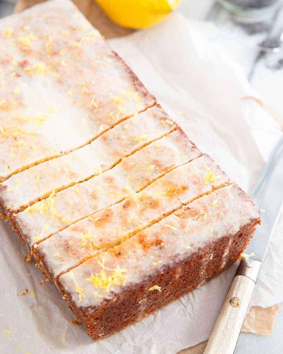 A rectangular loaf of lemon drizzle cake cut into six slices sits on parchment paper next to a knife.