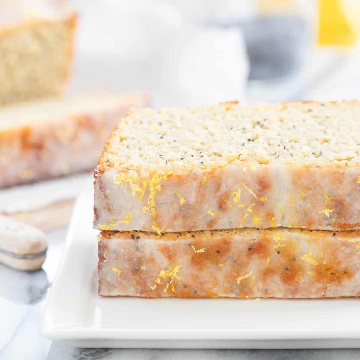 Two slices of lemon poppy seed bread on a white plate, showcasing a light drizzle of glaze and lemon zest on top. A knife is partially visible on the marble surface.