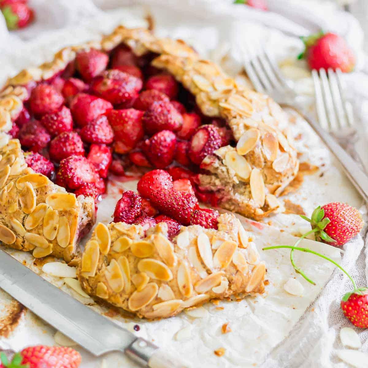 A rustic strawberry galette topped with sliced almonds sits on parchment paper, surrounded by fresh strawberries and two forks. A knife rests beside a cut slice, revealing the juicy strawberry filling inside the pastry.