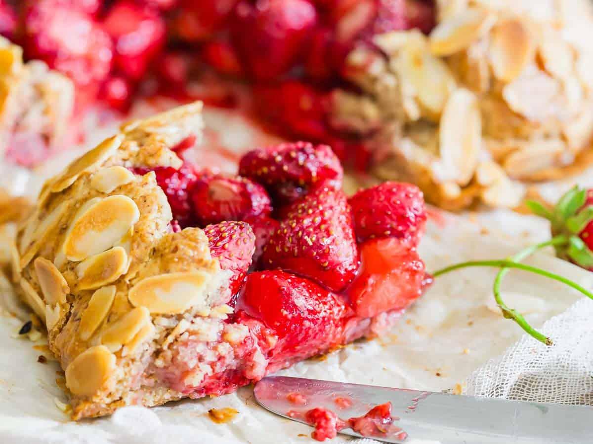 A close-up of a slice of strawberry galette showing juicy strawberries inside. The crust is golden brown and topped with sliced almonds. A knife rests nearby on the parchment paper.